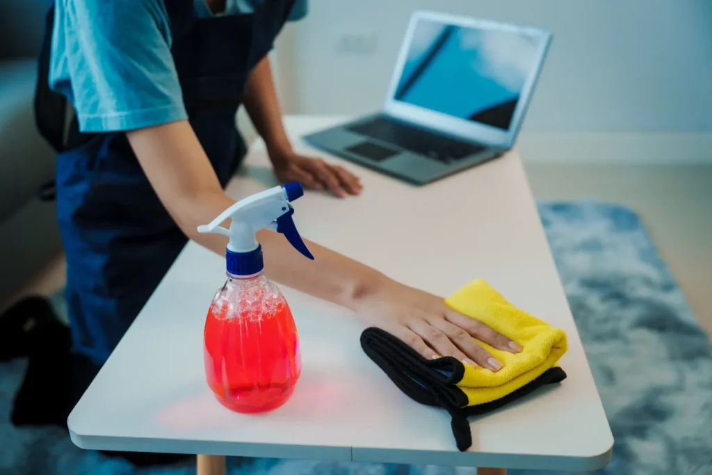 Persona limpiando un escritorio en oficina utilizando un limpiador multiusos para mantener la higiene y el orden en el entorno laboral.